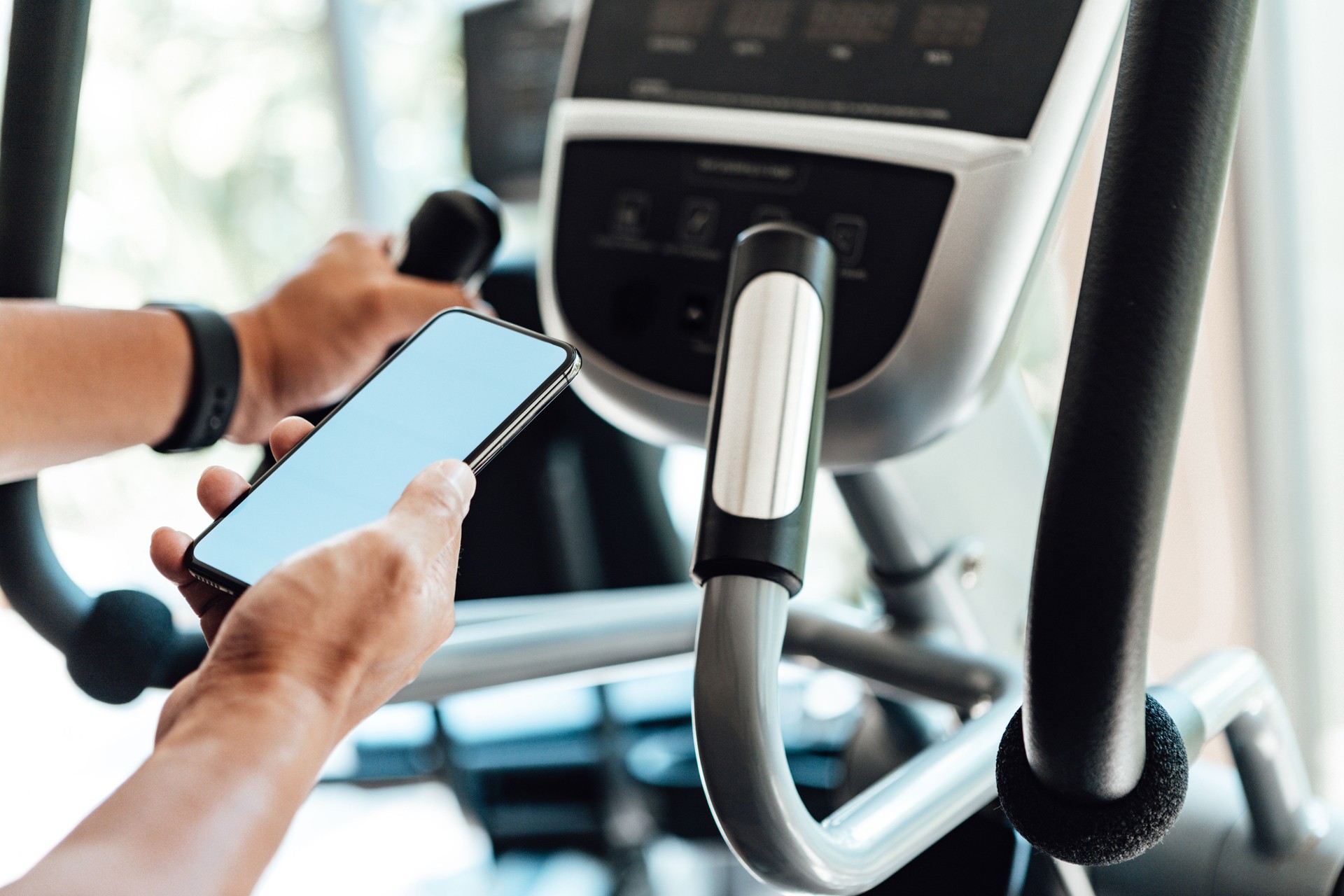 Determined Young Man Working Out On Elliptical Machine At Gym With Smart Phone